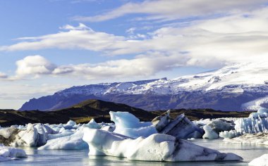Büyük bir su kütlesi buzla kaplıdır ve arka planda dağlar vardır. Gökyüzü açık ve mavi, sahne huzurlu ve huzurlu.