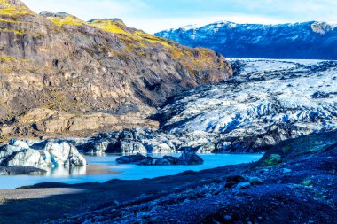 A beautiful landscape with a large body of water and Solheimajokull glacier, Iceland. The water is blue and the mountain is covered in moss clipart