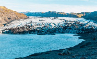 Solheimajokull buzulu ve arkasında karlı bir dağ olan güzel mavi bir göl. Bir çift sahilde durmuş manzaranın tadını çıkarıyor.