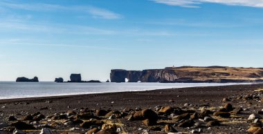 Arka planda büyük bir su kütlesi olan kayalık bir sahil. Gökyüzü açık ve mavidir, ve güneş parlıyor. Sahne huzurlu ve huzurlu. Dyrholaey, İzlanda