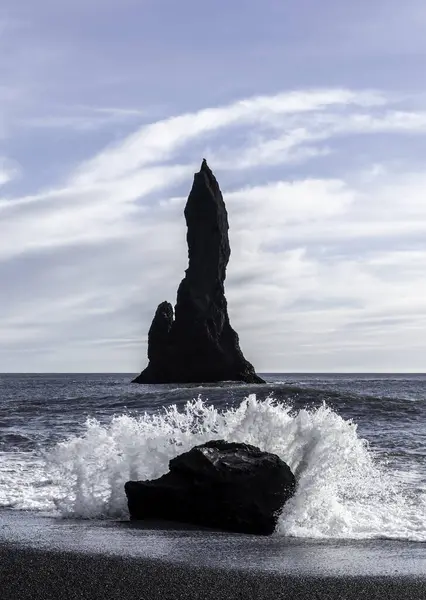 Reynisdrangar kulesi, Reynisfjara Kara Kum Sahili, İzlanda 'da. Okyanusta küçük bir dalganın yanında büyük bir kaya var. Kaya suyla çevrili ve gökyüzü berrak.