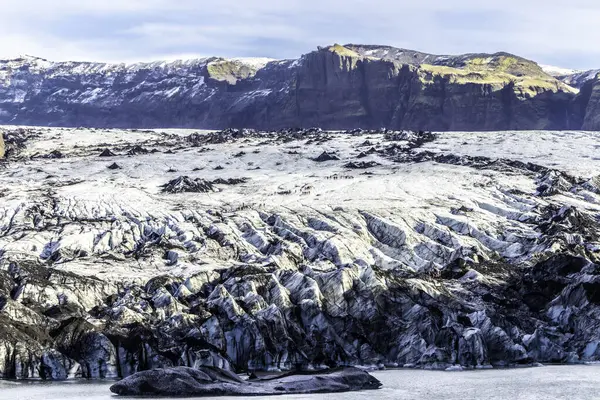 Önünde büyük bir buzul bulunan karlı bir dağ sırası. Solheimajokull Buzulu, İzlanda. Gökyüzü açık ve güneş parlıyor.