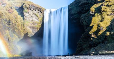 Arka planda gökkuşağı olan Skogafoss şelalesi. Şelale yemyeşil bir ormanla çevrilidir. İpek etkisi