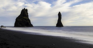 Reynisdrangar kulesi, Reynisfjara Kara Kum Sahili, İzlanda 'da. Önünde büyük kayalar olan kayalık bir sahil. Gökyüzü bulutlu ve su sakin. Sahne sakin ve huzurlu, kayalar ve su bir huzur hissi yaratıyor.