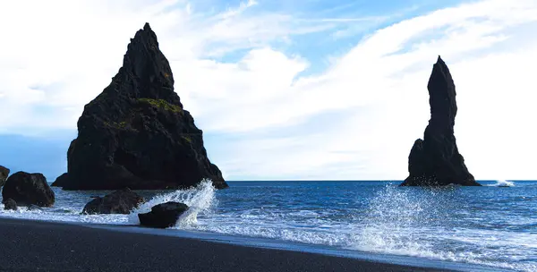 Reynisdrangar kulesi, Reynisfjara Kara Kum Sahili, İzlanda 'da. Ön planda büyük kayalar ve arka planda daha küçük bir kaya olan kayalık bir kıyı şeridi. Su sakin ve gökyüzü mavi.
