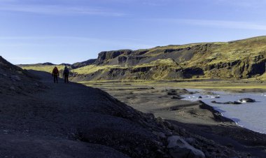 Bir su kütlesinin yanında kayalık bir yolda yürüyen iki kişi. Gökyüzü açık ve güneş ışıl ışıl parlıyor. Solheimajokull Buzulu, İzlanda