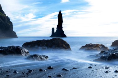 Reynisdrangar kulesi, Reynisfjara Kara Kum Sahili, İzlanda 'da. Uzakta büyük bir kayası olan kayalık bir sahil. Gökyüzü mavi ve su sakin. İpek etkisi