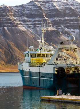 Büyük mavi bir tekne rıhtıma yanaştı. Tekne dağlarla çevrili ve su sakin.
