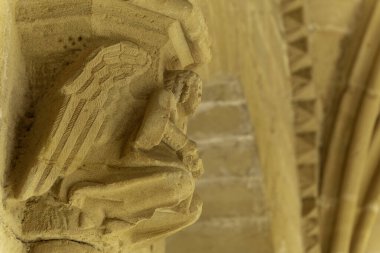 Elleri dizlerinde diz çökmüş bir melek heykeli. Heykel taştan yapılmıştır ve bir binanın üzerindedir. Ortaçağ mimarisi. Kilise Sanatının Ayrıntıları