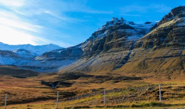 Tepesinde kar ve açık mavi gökyüzü olan bir dağ sırası. Dağın altındaki tarlayı çevreleyen bir ahşap ve tel çit var.