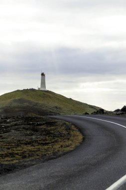 Uzakta deniz feneri olan bir yol. Yol boş ve gökyüzü bulutlu. Deniz feneri uzun ve beyaz