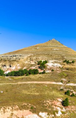 Arka planda mavi gökyüzü olan bir dağ. Dağ çimen ve ağaçlarla kaplıdır. Gökyüzü açık ve parlak