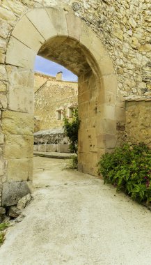 Önünde küçük bir bitki olan bir toprak yol, taş kemer deliğinden geçen taş bir duvarı geçer. Kemer yolu açık ve santral çiçeklerle dolu.