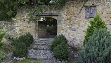 Taş kemerli taş bir bina ve ona doğru giden taş bir merdiven. Bina çalılar ve ağaçlarla çevrili.