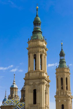 Yeşil kuleleri olan iki uzun, tuğladan kule. Bina mavi bir gökyüzü ile çevrilidir. El Pilar, Zaragoza Bazilikasının ayrıntıları