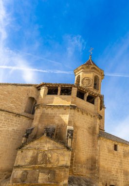 Üzerinde çan kulesi ve haç olan büyük ve eski bir kilise. Bina taştan yapılmıştır ve kırsal bir görünüme sahiptir. Gökyüzü açık ve mavi. Uzakta uçan bir uçak var.