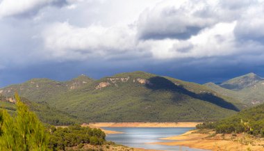 Önünde göl olan bir dağ sırası. Gökyüzü bulutlu ve dağlar ağaçlarla kaplı.