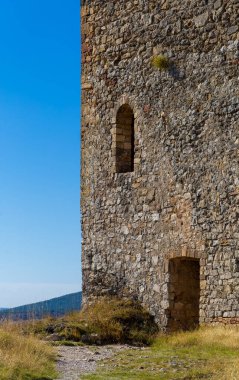 Arka planda mavi gökyüzü olan taş bir bina. Binanın küçük bir girişi ve büyük bir penceresi var.