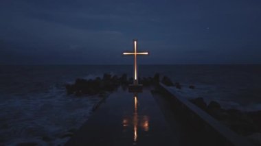 christian cross standing on pier in the stormy sea or ocean with dramatic sky at night