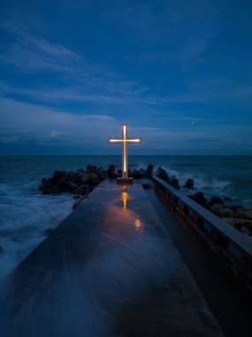 christian cross standing on pier in the stormy sea or ocean with dramatic sky at night