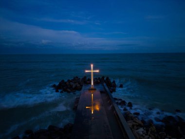 christian cross standing on pier in the stormy sea or ocean with dramatic sky at night
