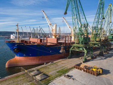 Arkaplanda işçilerin ve makinelerin görüldüğü yoğun bir limana yüklü veya tahılla indirilmiş bir gemi yanaşıyor.