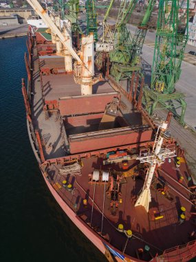 Arkaplanda işçilerin ve makinelerin görüldüğü yoğun bir limana yüklü veya tahılla indirilmiş bir gemi yanaşıyor.