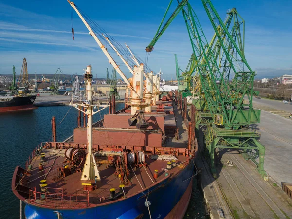 A bulk carrier ship loaded or downloaded with grain is docked at a busy port, with workers and machinery seen in the background.