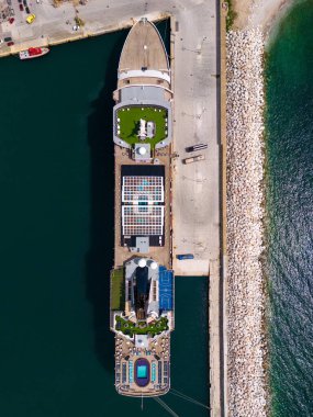 Çarpıcı hava manzarasını ele alan büyük bir yolcu gemisi hareketli limana kenetlenmiş, yüksek şehir manzarası ve ışıldayan mavi sularla çevrili. Hareketli sahne yolcularla dolu.