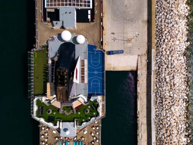 Çarpıcı hava manzarasını ele alan büyük bir yolcu gemisi hareketli limana kenetlenmiş, yüksek şehir manzarası ve ışıldayan mavi sularla çevrili. Hareketli sahne yolcularla dolu.