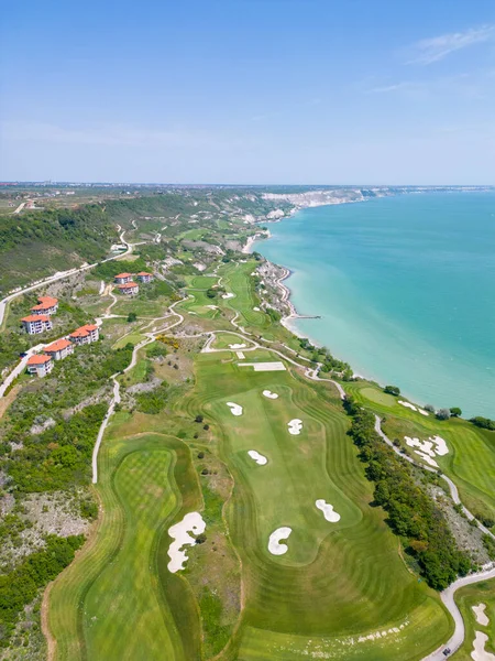 Aerial view of the golf course stretches out beneath, its lush green fairways complementing the breathtaking coastal panorama. Among the picturesque landscape, luxurious villas are strategically placed, offering a seamless fusion of elegance and scen