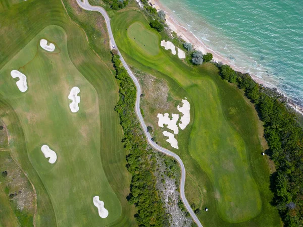 Aerial view of the golf course stretches out beneath, its lush green fairways complementing the breathtaking coastal panorama. Among the picturesque landscape, luxurious villas are strategically placed, offering a seamless fusion of elegance and scen