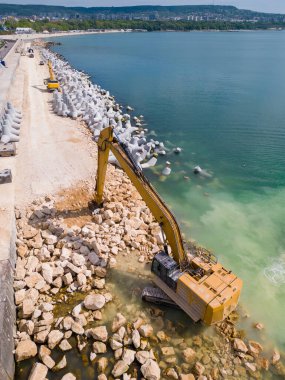 Bir kazı ustalıkla denizde bir rıhtım ya da dalgakıran inşa eder. Güçlü kolu kıyıdan uzanarak dalgaların arasında dirençli bir yapı oluşturur. Havadan üst görünüm