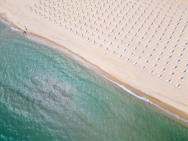 Kumlu bir sahil sahil boyunca uzanır, boş güneş pansiyonlarıyla süslenir. Havadan bakıldığında, sakin bir manzara gözler önüne seriliyor. Gök mavisi deniz, altın kıyı şeridiyle buluşuyor. Boş güneşlikler.