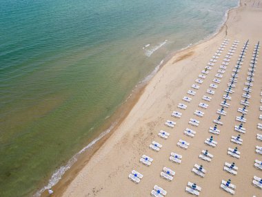 Kumlu bir sahil sahil boyunca uzanır, boş güneş pansiyonlarıyla süslenir. Havadan bakıldığında, sakin bir manzara gözler önüne seriliyor. Gök mavisi deniz, altın kıyı şeridiyle buluşuyor. Boş güneşlikler.
