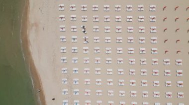 Kumlu bir sahil sahil boyunca uzanır, boş güneş pansiyonlarıyla süslenir. Havadan bakıldığında, sakin bir manzara gözler önüne seriliyor. Gök mavisi deniz, altın kıyı şeridiyle buluşuyor. Boş güneşlikler.