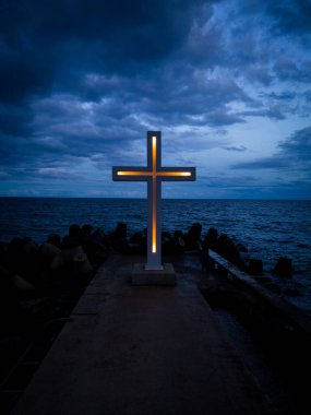 Büyük bir Hıristiyan haçı, bir rıhtımın kenarında, yukarıdan görülen dramatik bir gökyüzü ve denize karşı duruyor..