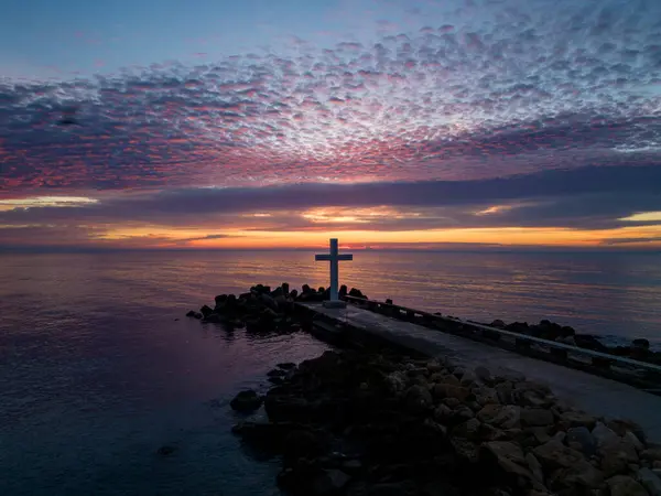 A flight around a Christian cross early in the morning at sunrise. The large cross stands on the edge of a breakwater on the sea coast.