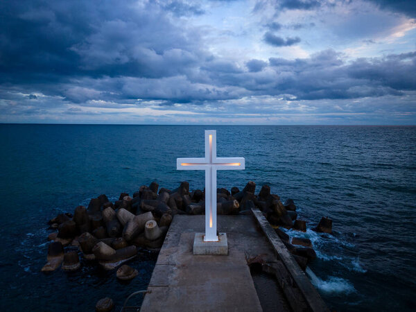 A large Christian cross stands at the edge of a pier against a dramatic sky and sea, seen from above.