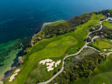 Okyanusun arka planına kurulmuş yeşil çayırları ve mavi suları gösteren bir golf sahasının genel manzarası..