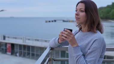 Elinde kahve fincanı olan genç bir kadın romantik bir şekilde denizin kenarına bakıyor..