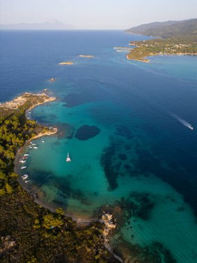 Yunanistan 'ın Sithonia Yarımadası' nda kristal berrak sularla çevrili plajları ve kayaları olan bir adanın havadan görünüşü. Yatlar ve tekneler turkuaz sularda demirliyor. Yakındaki adalar ve resifler görülebilir..