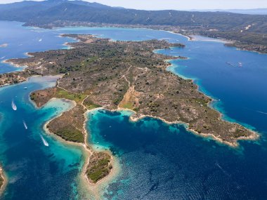 Aerial view of an island on Sithonia Peninsula, Greece, with beaches and rocks, surrounded by crystal-clear waters. Yachts and boats anchor in turquoise waters. Nearby islets and reefs visible. clipart