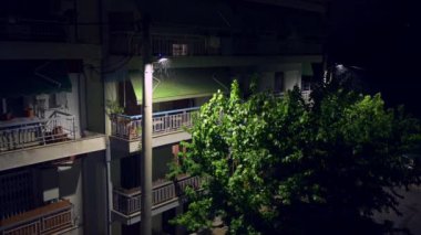 Şehirde yağmurlu bir gecenin sükuneti. Sokak lambaları ve yemyeşil yapraklarla aydınlatılmış bir konut binasının altını çizmek.