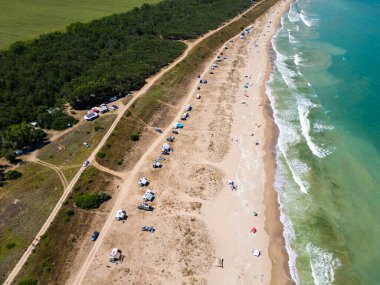 Sahil boyunca dağınık çadırları ve şemsiyeleri olan kumlu bir sahil manzarası. Dalgalar yavaşça kıyıya vurur, okyanus kıyısında huzurlu ve rahatlatıcı bir yaz gününü yakalar..