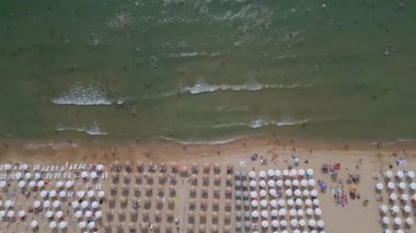 Kalabalık bir plajın, okyanusun nazik dalgalarına karşı dizilmiş şemsiyeli ve güneşli videoları. Hava görüntüsü, bir yaz gününün canlı atmosferini yakalar. Bulgaristan.