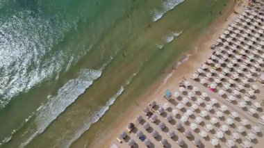 Kalabalık bir plajın, okyanusun nazik dalgalarına karşı dizilmiş şemsiyeli ve güneşli videoları. Hava görüntüsü, bir yaz gününün canlı atmosferini yakalar. Bulgaristan.