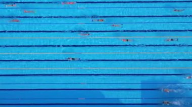 Yüzücülerin açık mavi bir havuzda, yukarıdan çekilen drone videosu. Yukarıdan aşağıya bakış, yüzücülerin ritmik hareketlerini ve suyun canlı, ferahlatıcı atmosferini vurgular..