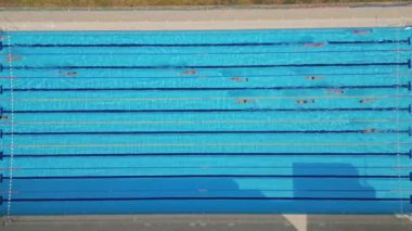 Yüzücülerin açık mavi bir havuzda, yukarıdan çekilen drone videosu. Yukarıdan aşağıya bakış, yüzücülerin ritmik hareketlerini ve suyun canlı, ferahlatıcı atmosferini vurgular..