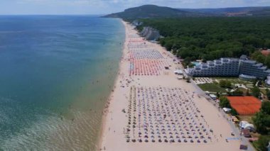 Sırayla dizilmiş şemsiyeler, güneş banyosu ve su aktiviteleriyle dolu geniş bir plajın insansız hava aracı videosu. Hava manzarası, deniz kıyısındaki tatil beldesindeki bir yaz gününün canlı atmosferini yakalar. Albena, Bulgaristan.